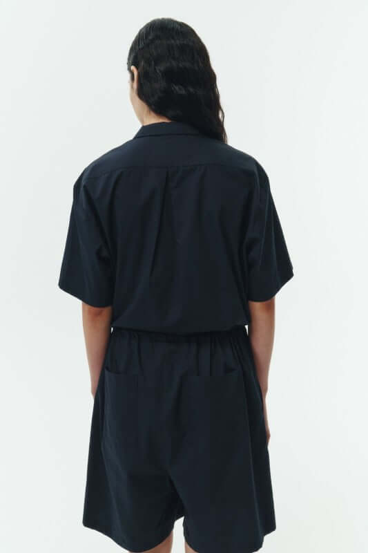 Back view of a woman wearing the Emma Solid Jumpsuit in black, showcasing its relaxed fit and camp collar design.